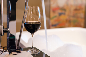 A glass of red wine with book and spectacles put on table beside bathtub that have white bubble. Relaxing at home concept.