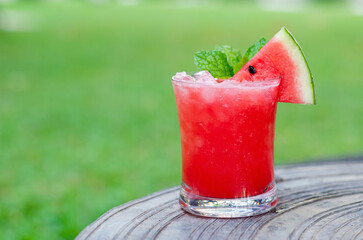 A glass of watermelon smoothie puts on table with blurred green yard background for summer drink concept.