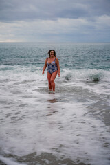 A plump woman in a bathing suit enters the water during the surf. Alone on the beach, Gray sky in the clouds, swimming in winter.