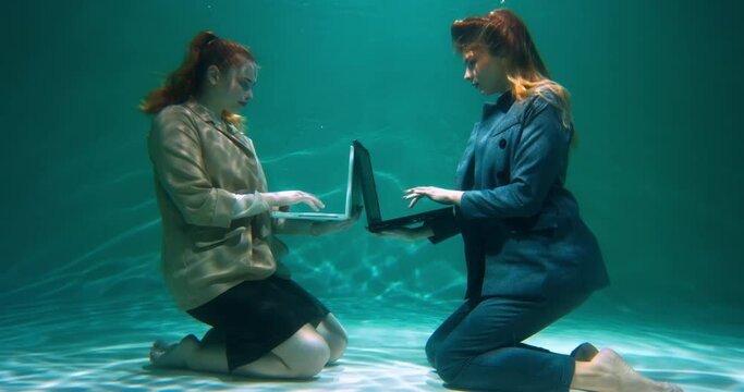 Two young happy busy female managers in suits work online writing code underwater, closing laptops rising up slow motion