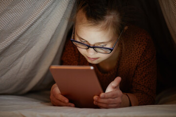 Portrait of young girl with down syndrome using smartphone in bed hiding under covers, digital...