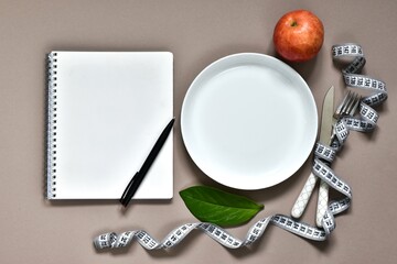 Composition with cutlery, plate, measuring tape, paper notebook with text DIET PLAN and green leaves on beige background. Flat lay, top view with copy space
