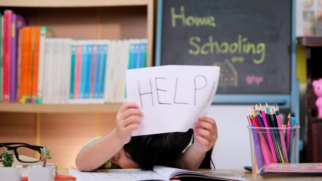 Asian little girl feeling sad and tired sitting at the table with many books and holding a paper with word Help. Learning difficulties. Home schooling.