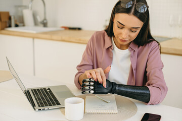 Good-looking stylish young girl adjusting settings of her metal bionic prosthetic arm touching sensory panel on upper side of artificial hand, sitting at kitchen table in front of laptop, working