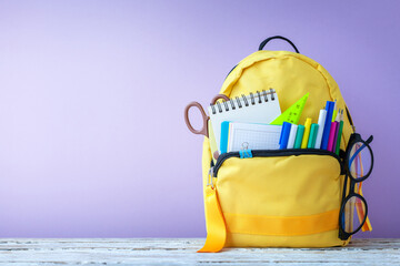 Full yellow school backpack with stationery and notes, pens, glasses on table on purple background. Concept back to school.