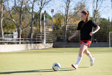 Beautiful girl joyfully playing football. Young girl in sport uniform training walking on field on sunny day, looking at ball and preparing to kick. Sport activity, active rest and hobby concept