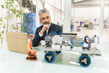 Businessman sitting with hand on chin looking at model of robotic vehicle in factory