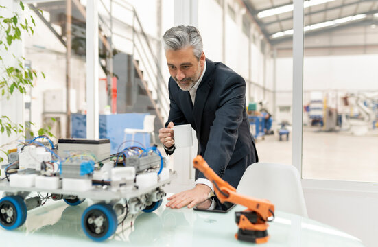 Businessman holding coffee cup examining robotic vehicle model standing at desk