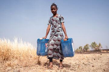 Grim and tired looking African girl with two water cans in her hands walking on a dirt road in a...