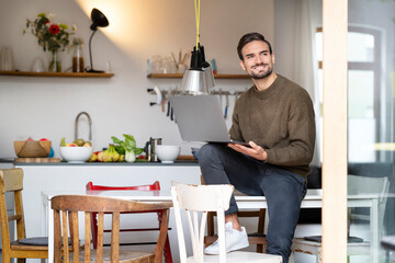 Smiling freelancer with laptop sitting on table in kitchen at home