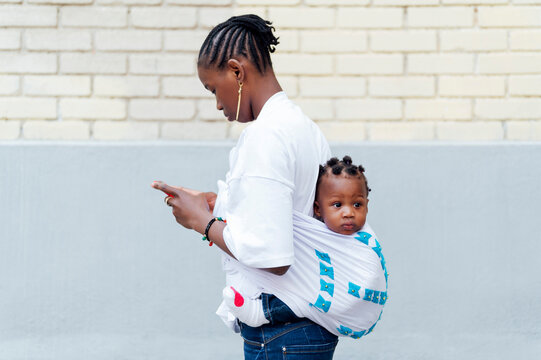 Young woman using smart phone carrying daughter in kanga baby carrier