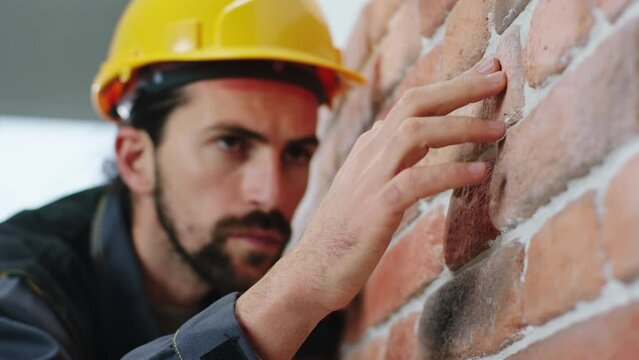 In front of the camera closeup charismatic and good looking man constructor worker analysing the structure of red brick from the new wall built he wearing safety helmet and uniform