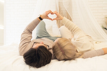 Young married Caucasian people lying on the bed spending free time together showing heart gesture...