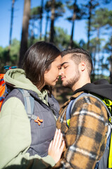 Loving couple of young hikers. Caucasian man with beard and woman in cap with big backpacks touching with noses tenderly in forest. Hobby, nature, love concept