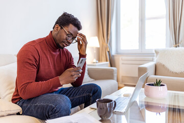 Fototapeta premium African man sit on couch using smartphone shocked by reading online news, surprised male get breaking message or text on mobile phone, confused guy holding cell see warning notification