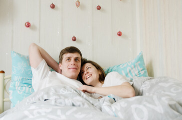 Happy couple lying in bed in cozy home, drinking coffee. Young man in a t-shirt and a cute woman....
