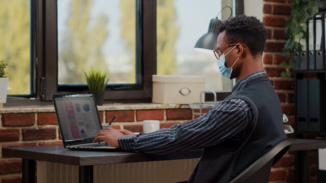African american worker analyzing commercial growth on laptop in startup office. Company employee wearing face mask and working on marketing development with sales report on computer.
