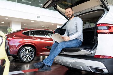 a buyer studying a booklet in a showroom of a car dealership sitting in the trunk of a crossover looking at the camera with a smile - obrazy, fototapety, plakaty