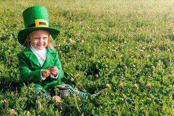 St. Patrick's Day. A girl in a green suit is sitting in a large clearing of clover, holding a pot...