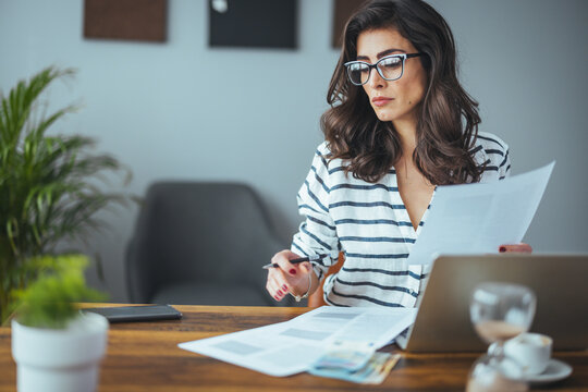 Indoor shot of casually dressed young woman holding papers in her hands, calculating family budget, trying to save some money , having stressed and concentrated look
