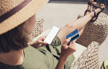 Young woman on vacation holding credit card and using mobile phone with blank empty mockup screen,...