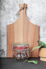 Kitchen background with spices. Black pepper, peppercorns in a glass jar and bay leaf in a wooden mortar on the kitchen table against the background of a cutting board. Kitchen utensils front view