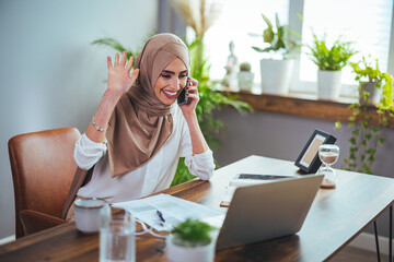 Smiling Muslim woman talking on the phone, studying online with a Skype teacher, happy young Muslim...