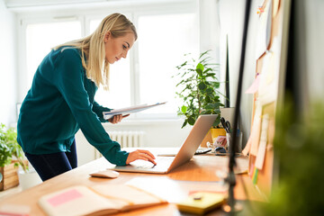 Young woman standing by a desk using laptop holding documents working from home office 