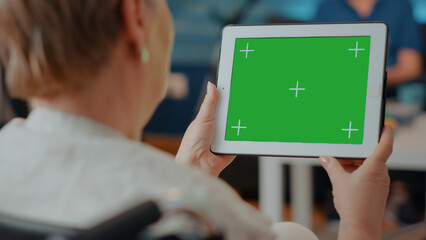 Old woman with disability holding green screen on digital tablet. Senior person sitting in...