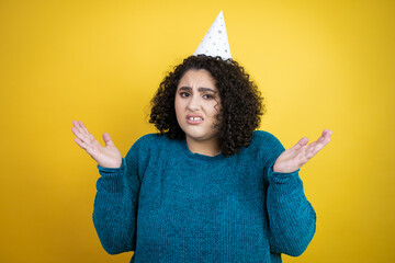 Young beautiful woman wearing a birthday hat over isolated yellow background clueless and confused expression with arms and hands raised - obrazy, fototapety, plakaty
