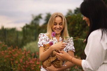 A young mother with a teenage daughter playing with a ginger cat in the garden. Mom and her daughter teenage girl are playing , smiling and hugging. Family holiday and togetherness.