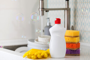 A white bottle with dishwashing gel, sponges and rubber gloves on the background of a sink and flying soap bubbles.