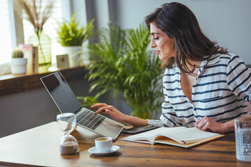 A shot of a young woman using a laptop and going through paperwork while working from home. A woman who uses a laptop while sitting at home. The concept of working from home