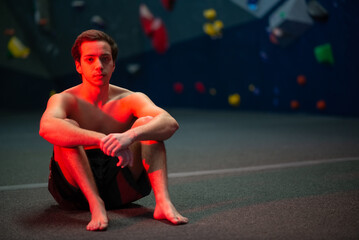 Fototapeta premium Young sportive man resting after training in gym. Sportsman in dark shorts sitting on floor and looking at camera. Sport, hobby, wellbeing concept