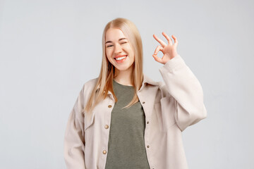 Smiling young pretty woman gesturing OK, showing okay sign and looking at the camera over grey...