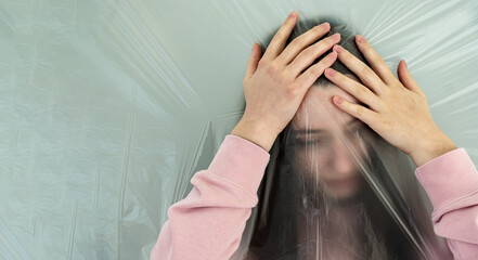 young woman with distorted face behind plastic film grabs her head, because of headache, the...