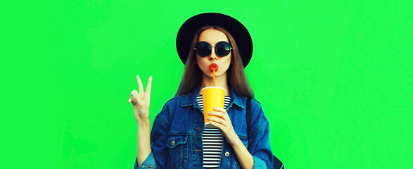 Portrait of young woman model drinking juice wearing black round hat, denim jacket on green background