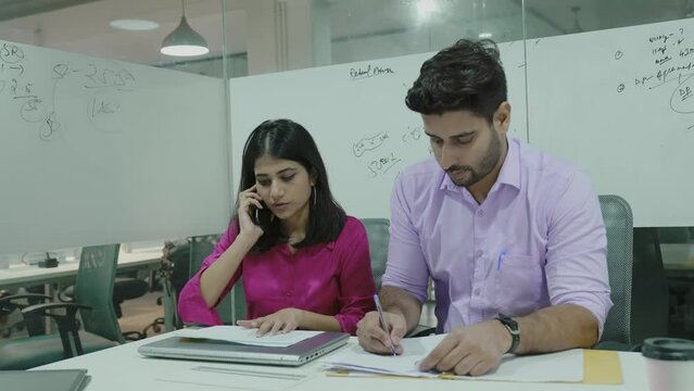 Young Indian woman talking to manager on phone call about business project while man checking rate charts on papers and taking notes Colleagues working together to design marketing presentation.