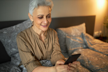 Good-looking senior Caucasian woman in brown striped blouse holding smartphone in hands watching videos on social networks, smiling, having stylish haircut, and natural daily make-up, sitting on bed