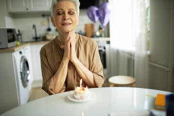 Obraz na płótnie Canvas Excited enthusiastic charming elderly woman in brown keeping eyes closed, hands folded in praying position, making wish sitting in front of cupcake with lit candle against cozy kitchen and balloons