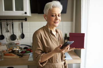 Caucasian senior lady standing against kitchen tabletop with vegetables and utensils, in front of window with smartphone and present box in hands, dialing number, wearing white earphones