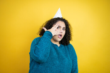 Young beautiful woman wearing a birthday hat over isolated yellow background peeking in shock covering face and eyes with hand, looking through fingers with embarrassed expression