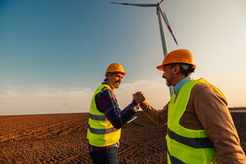 Two engineers shaking hands on site at wind turbines field or farm,