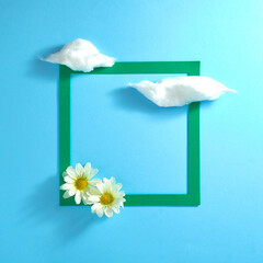 Fluffy white clouds, two white flowers, and a green frame on a pastel blue background. Creative. Text space. Top view. Minimal style.