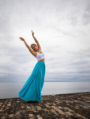 Beautiful woman practicing ballet pose. Young ballerina wearing long blue skirt and dancing. Outdoor ballet practice. Slim body. Cloudy sky background. Copy space. Bali