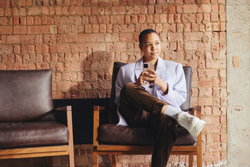 Portrait of multiracial LGBTQ mid adult woman using smartphone on retro chair in front of exposed brick wall and looking away