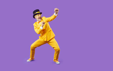 Fototapeta na wymiar Funny senior man fighting imaginary enemy. Full body shot of happy crazy silly old man wearing yellow suit and top hat fighting invisible opponent and having fun isolated on solid purple background