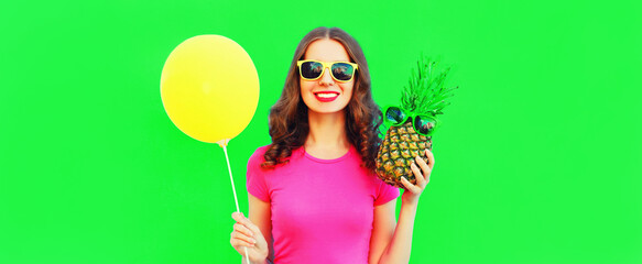 Summer portrait of happy smiling young woman with pineapple and yellow balloon on green background