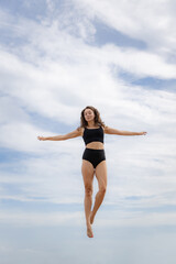 Fototapeta na wymiar Attractive young woman jumping over cloudy blue sky. Caucasian woman wearing black sportswear. Fitness, wellness concept. Outdoor activity. Copy space. Sky background. Bali