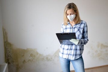 pretty young woman standing in front of wall with mold with tablet and mouth nose mask